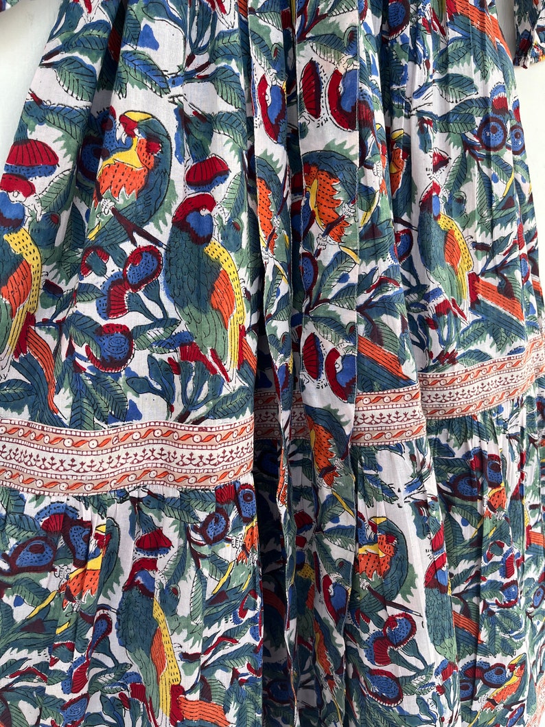 May include: A white cotton dress with a colorful floral print featuring parrots, leaves, and berries. The dress has a tiered skirt and a fitted bodice.
