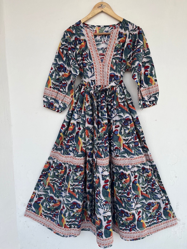 May include: A white maxi dress with a colorful bird and floral print. The dress has a V-neckline, 3/4 sleeves, and a tie waist. The dress is made of cotton and has a tiered skirt.
