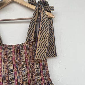 May include: A brown and blue patterned dress with a smocked bodice and a gathered skirt. The dress has thin straps that tie at the shoulders. The straps are made of the same fabric as the dress and have a zig-zag pattern.