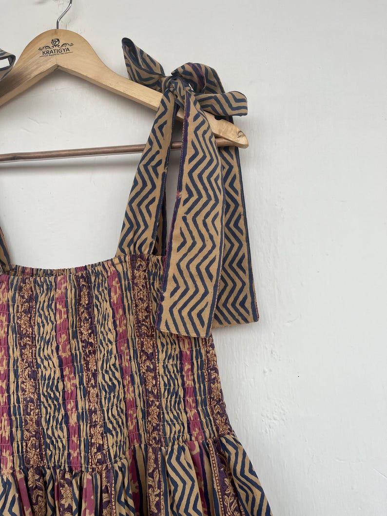 May include: A brown and blue patterned dress with a smocked bodice and a gathered skirt. The dress has thin straps that tie at the shoulders. The straps are made of the same fabric as the dress and have a zig-zag pattern.