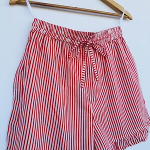 May include: Red and white striped shorts with a drawstring waist and ruffled hem. The shorts are hanging on a wooden hanger against a white background. The vertical stripes create a classic, summery look.