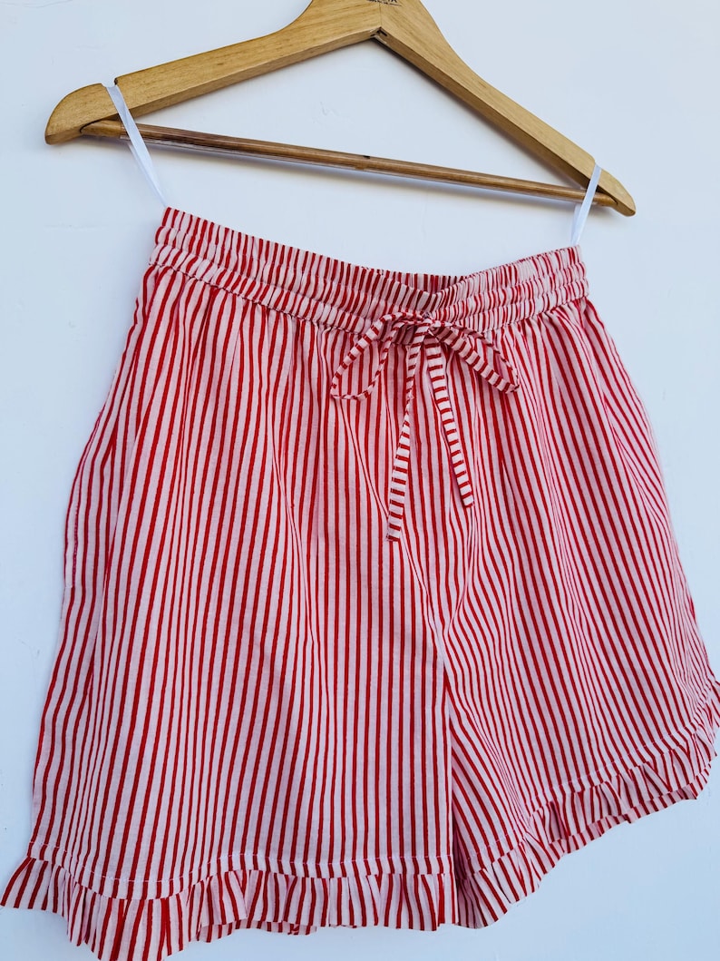 May include: Red and white striped shorts with a drawstring waist and ruffled hem. The shorts are hanging on a wooden hanger against a white background. The vertical stripes create a classic, summery look.