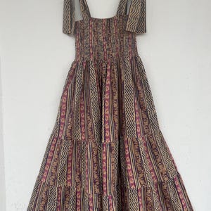May include: A tiered maxi dress with a brown and pink geometric print. The dress has a square neckline and thin straps that tie at the shoulders.