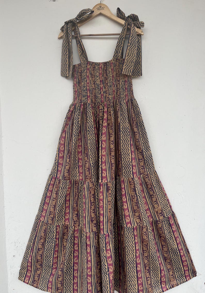 May include: A tiered maxi dress with a brown and pink geometric print. The dress has a square neckline and thin straps that tie at the shoulders.