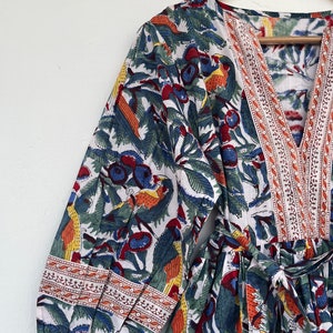 May include: A white cotton dress with a colorful floral print featuring birds and berries. The dress has a V-neckline, long sleeves, and a tie waist. The sleeves and neckline are trimmed with a red, white, and blue embroidered border.