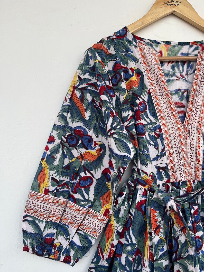 May include: A white cotton dress with a colorful floral print featuring birds and berries. The dress has a V-neckline, long sleeves, and a tie waist. The sleeves and neckline are trimmed with a red, white, and blue embroidered border.