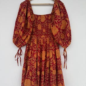 May include: A red and orange floral print dress with a square neckline and puffed sleeves. The dress has a tiered skirt and tie details on the sleeves. The dress is hanging on a wooden hanger.