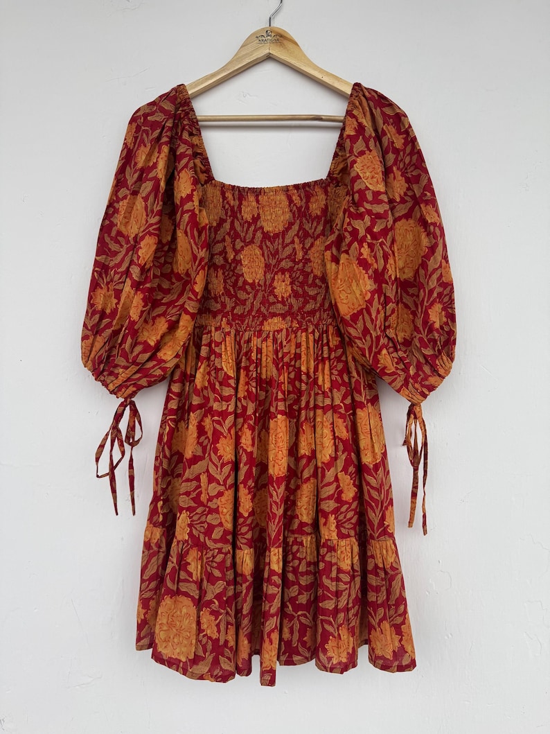 May include: A red and orange floral print dress with a square neckline and puffed sleeves. The dress has a tiered skirt and tie details on the sleeves. The dress is hanging on a wooden hanger.