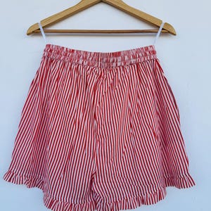 May include: Red and white striped shorts with a ruffled hem, hanging on a wooden hanger. The shorts have an elastic waistband and a vertical stripe pattern. The garment is displayed against a white background.