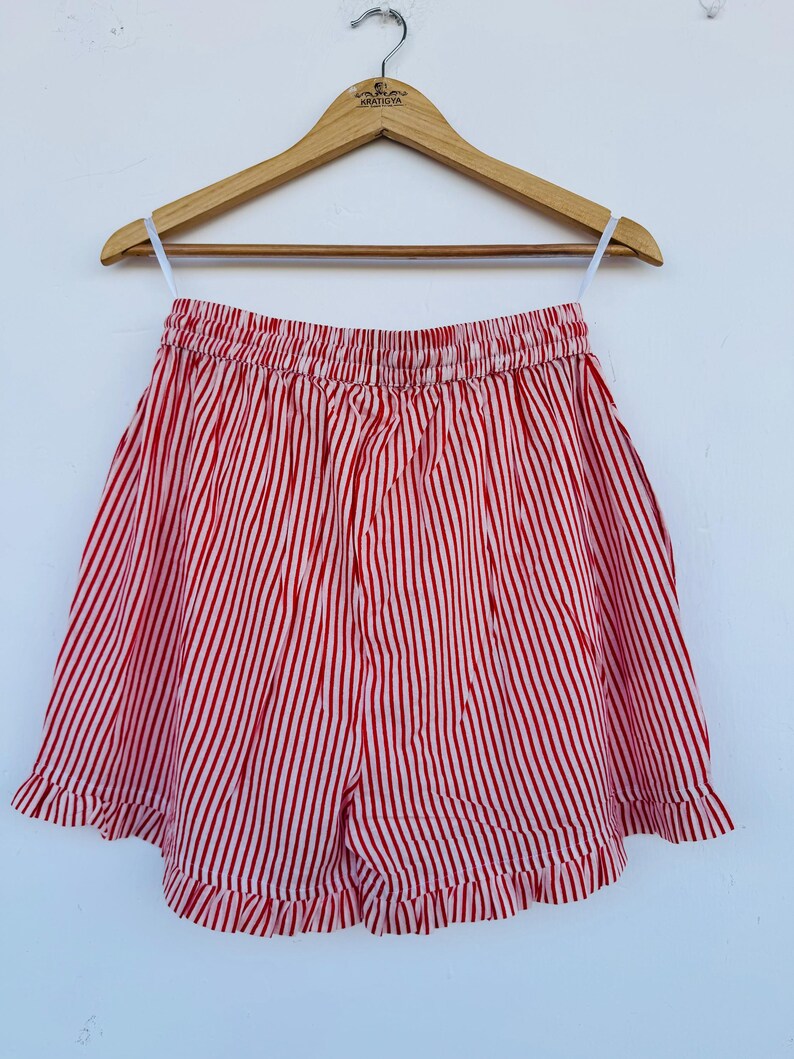 May include: Red and white striped shorts with a ruffled hem, hanging on a wooden hanger. The shorts have an elastic waistband and a vertical stripe pattern. The garment is displayed against a white background.