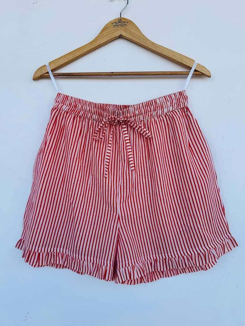 May include: Red and white striped shorts with a drawstring waist and ruffled hem. The shorts are hanging on a wooden hanger against a white background. The vertical stripes create a classic, summery look.