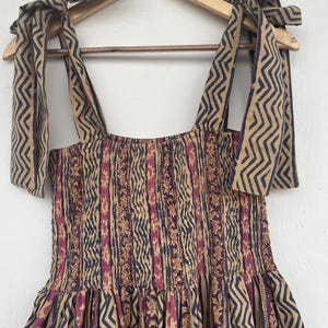 May include: A brown and pink patterned dress with a smocked bodice and tie straps. The straps are made of the same fabric as the dress and have a zig-zag pattern. The dress is hanging on a wooden hanger with the word "KRATIGYA" printed on it.