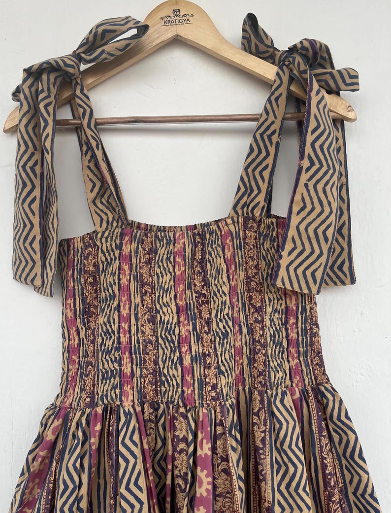 May include: A brown and pink patterned dress with a smocked bodice and tie straps. The straps are made of the same fabric as the dress and have a zig-zag pattern. The dress is hanging on a wooden hanger with the word "KRATIGYA" printed on it.