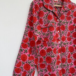 May include: A pink long-sleeved shirt with a red and green floral print. The shirt has a button-up front and a collar.