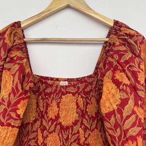 May include: A red and orange floral print dress with a square neckline and puffed sleeves. The dress is hanging on a wooden hanger with the brand name "KRATIGYA" visible. The fabric has a repeating pattern of large flowers and leaves.