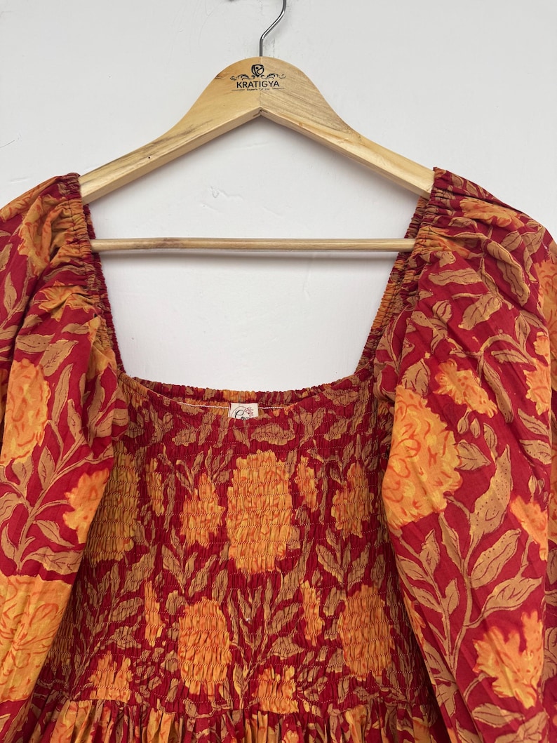 May include: A red and orange floral print dress with a square neckline and puffed sleeves. The dress is hanging on a wooden hanger with the brand name "KRATIGYA" visible. The fabric has a repeating pattern of large flowers and leaves.