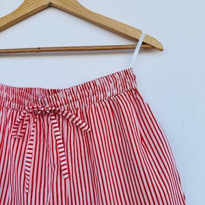 May include: Red and white striped shorts hanging on a wooden hanger. The shorts have a drawstring waist and are made of a lightweight fabric. The vertical stripes are evenly spaced and create a classic, summery look.
