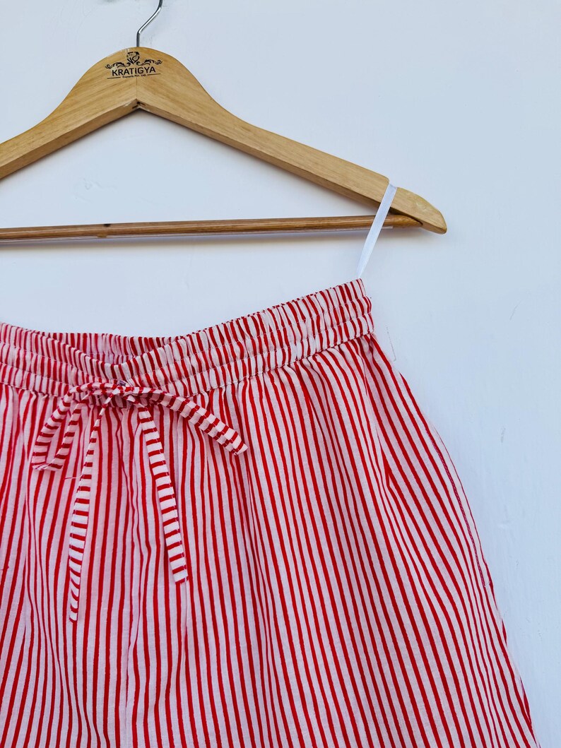 May include: Red and white striped shorts hanging on a wooden hanger. The shorts have a drawstring waist and are made of a lightweight fabric. The vertical stripes are evenly spaced and create a classic, summery look.