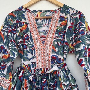 May include: A white cotton dress with a floral print featuring blue, red, orange, and green colors. The dress has a V-neckline with a decorative trim and a tie at the waist.