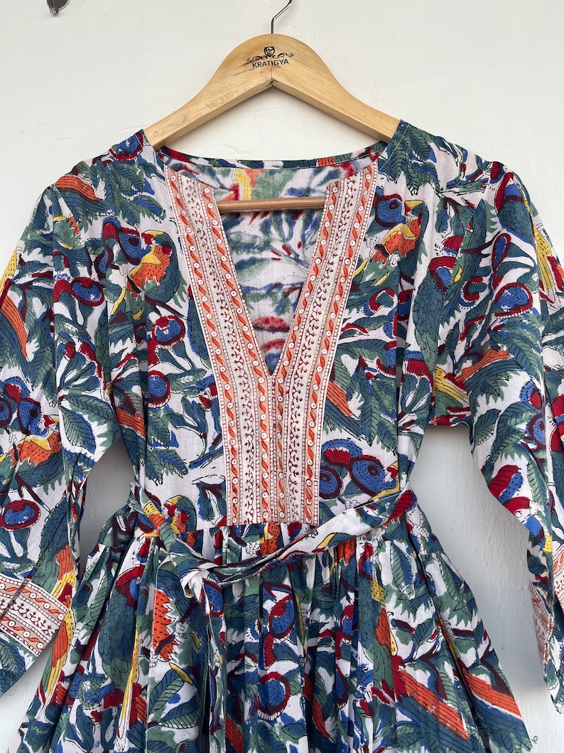 May include: A white cotton dress with a floral print featuring blue, red, orange, and green colors. The dress has a V-neckline with a decorative trim and a tie at the waist.