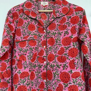 May include: A pink and red long-sleeved shirt with a floral print. The shirt has a button-up front and a collar. The print features red flowers with green leaves on a pink background.