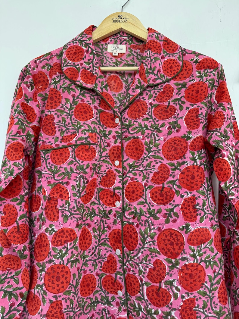 May include: A pink and red long-sleeved shirt with a floral print. The shirt has a button-up front and a collar. The print features red flowers with green leaves on a pink background.