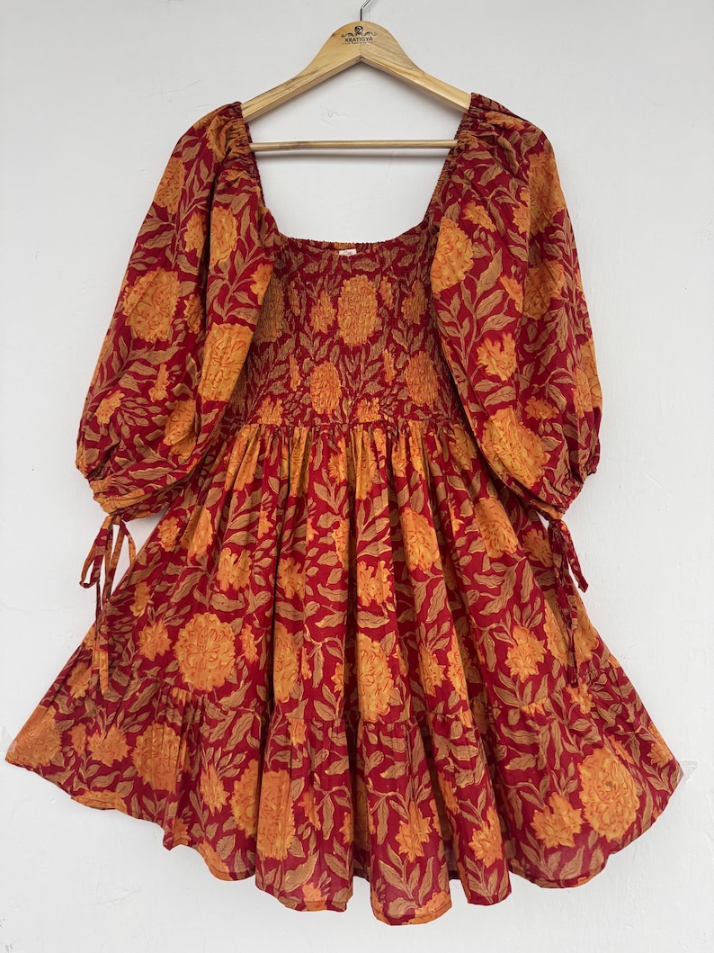 May include: A red and orange floral print dress with a square neckline and long, puffed sleeves. The dress features a smocked bodice and a tiered skirt. The sleeves have tie details at the cuffs. The dress is hanging on a wooden hanger.