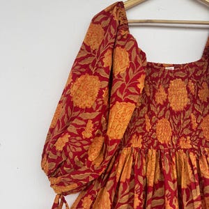 May include: A red and orange floral print dress with a square neckline and puffed sleeves. The dress is hanging on a wooden hanger. The fabric features a repeating pattern of large flowers and leaves.