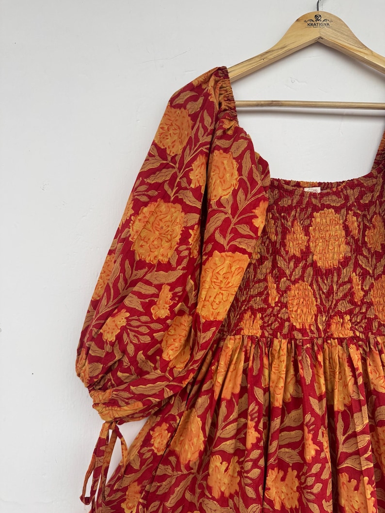 May include: A red and orange floral print dress with a square neckline and puffed sleeves. The dress is hanging on a wooden hanger. The fabric features a repeating pattern of large flowers and leaves.
