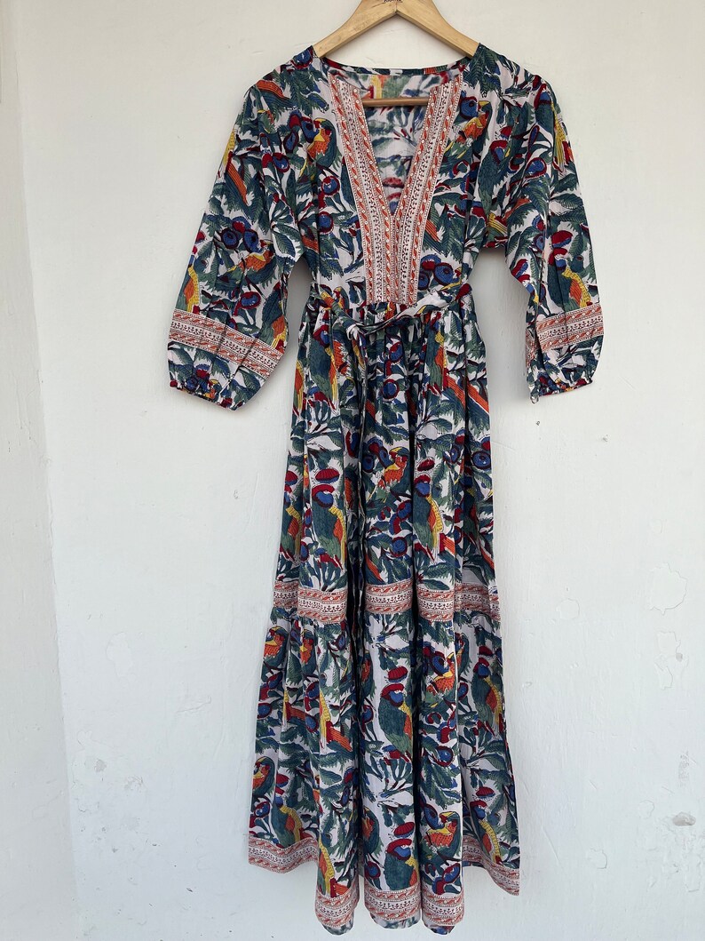 May include: A white maxi dress with a colorful floral print featuring birds and leaves. The dress has a V-neckline, 3/4 sleeves, and a tie waist.