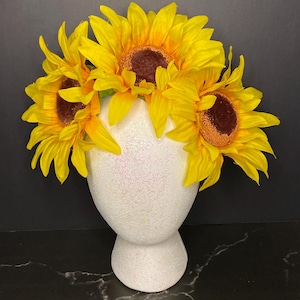 May include: A decorative head form adorned with bright yellow sunflowers. The sunflowers have brown centers and vibrant yellow petals. The head form is white with a glittery texture, set against a dark background.