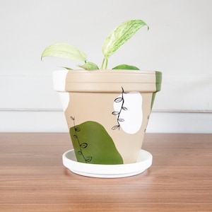 Puede incluir: Una maceta de terracota beige con un borde blanco y un diseño abstracto verde y blanco. La maceta tiene un platillo blanco y está llena de una planta verde con variegación blanca.