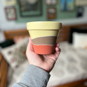 Könnte beinhalten: Ein handbemalter Keramik-Blumentopf mit einem geschichteten Design. Der obere Bereich ist gelb, gefolgt von einem braunen Band und einer korallenfarbenen Basis. Der Topf wird vor einem verschwommenen Hintergrund mit gerahmten Kunstwerken gehalten.