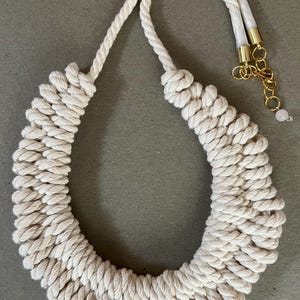 Puede incluir: Collar de cuerda color crema con un diseño grueso y tejido. El collar presenta una sección central grande en forma de U y una cuerda más fina para el cuello. Cadena dorada y una pequeña cuenta rosa completan el diseño.