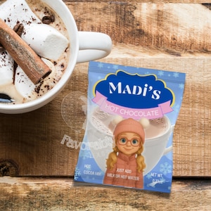 Peut inclure: Une tasse de chocolat chaud avec des guimauves et un bâton de cannelle à côté d'un sachet de chocolat chaud Madi's. Le sachet présente une fille de dessin animé et le texte "Just add milk or hot water". Le poids net du sachet est de 20.69g.