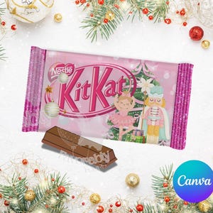 May include: A pink Kit Kat candy bar with a Christmas-themed design featuring a ballerina and a nutcracker. The candy bar is partially unwrapped, revealing the chocolate. The background includes Christmas ornaments and greenery.
