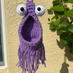 May include: Purple crocheted hanging planter with a large opening and fringe. The planter has two large white googly eyes with black pupils.