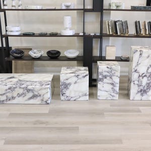 May include: Four white marble pedestal tables with purple veining. The tables are arranged in a row on a light wood floor.