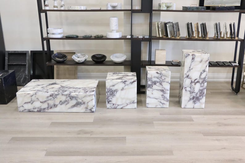 May include: Four white marble pedestal tables with purple veining. The tables are arranged in a row on a light wood floor.