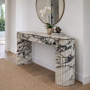 Puede incluir: Una consola de mármol con un patrón veteado en blanco y negro. La mesa tiene un cajón y patas acanaladas. Un espejo redondo dorado cuelga encima de la mesa, con una orquídea en maceta y objetos decorativos.