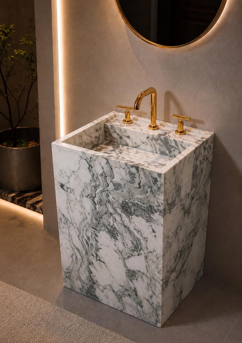 Puede incluir: Un lavabo rectangular independiente hecho de m&aacute;rmol blanco con vetas grises prominentes. El lavabo cuenta con un grifo de oro pulido con dos manijas en forma de T a juego montadas en el borde trasero plano de la estructura de piedra.