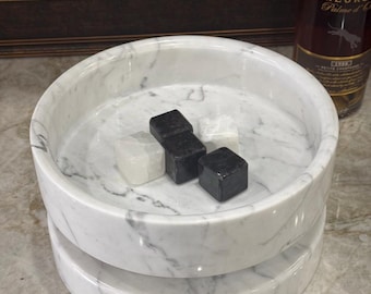 May include: A white marble serving tray with gray veining, featuring a two-tiered design. The tray holds three square whiskey stones, two black and one white. A bottle of cognac is visible in the background.