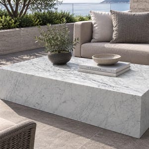 May include: A rectangular white marble coffee table with grey veining. On the table sits a small dark grey pot containing a green plant, a shallow beige bowl, and a stack of two books with the word The Kinfolk printed on the top cover.