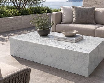May include: A rectangular white marble coffee table with grey veining. On the table sits a small dark grey pot containing a green plant, a shallow beige bowl, and a stack of two books with the word The Kinfolk printed on the top cover.