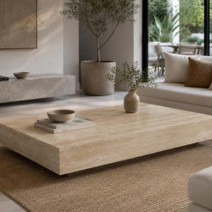 Pode incluir: Uma mesa de centro retangular e baixa feita de pedra travertino bege com uma textura porosa natural. A mesa apresenta um design de bloco espesso e sólido, colocada sobre um tapete castanho texturizado, com uma pequena taça e livros em cima.