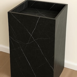 Puede incluir: Un pedestal rectangular alto con acabado negro y vetas blancas en toda la superficie. La estructura presenta un dise&ntilde;o hueco con la parte superior abierta y una base plana, mostrando una silueta geom&eacute;trica limpia con bordes definidos.