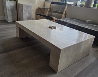 May include: A rectangular coffee table constructed from beige travertine stone with a smooth, polished surface and a solid, block-style base. A small, light-colored wooden starfish ornament is placed in the center of the table top.
