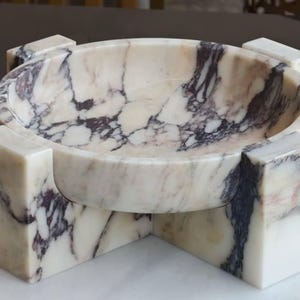 May include: A marble bowl with a circular basin, supported by four rectangular legs. The bowl and legs are white with dark purple veining. The bowl is a decorative item.