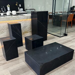Könnte beinhalten: Eine Sammlung von schwarzen Marmormöbeln, darunter ein rechteckiger Couchtisch, zwei quadratische Beistelltische und zwei hohe Sockel. Der Marmor hat weiße Adern, und die Stücke sind auf einem hellen Holzboden angeordnet.