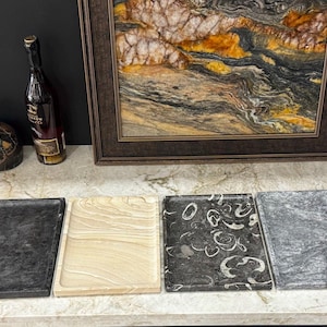 May include: Four rectangular stone trays in varying colors and patterns. The trays include a dark gray, a light beige with wood grain, a dark gray with white circular patterns, and a light gray with darker gray veining. A framed artwork and a bottle are in the background.